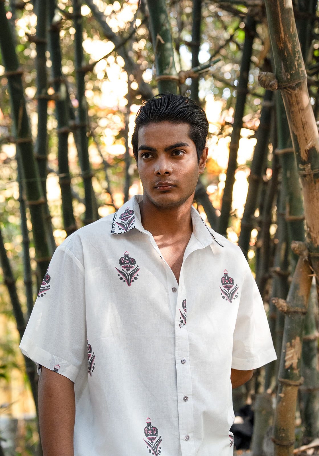 A man wearing a white short-sleeve button-up shirt with embroidered crown motifs, standing in front of a bamboo forest.